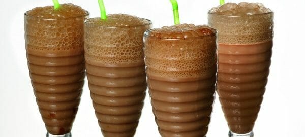 Four soda fountain glasses filled with a mix of chocolate milk and seltzer water, with green straws.