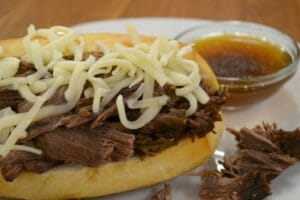 Close-up of a French Dip sandwich with shredded cheese on a white plate, with a side bowl of au jus.