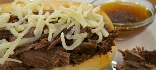 Close-up of a French Dip sandwich with shredded cheese on a white plate, with a side bowl of au jus.