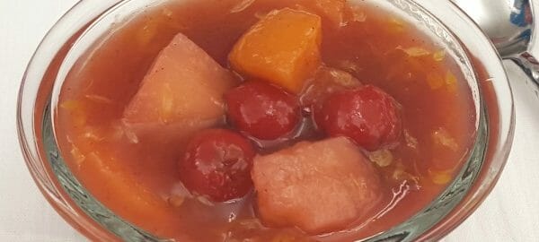 A clear ramekin filled with a serving of fruit soup, including apricots, pineapple, and cherries.