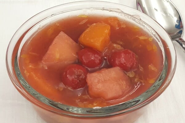 A clear ramekin filled with a serving of fruit soup, including apricots, pineapple, and cherries.