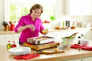 A woman in a fuschia blouse in a kitchen, sprinkling shredded cheese on a pan of lasagna, with a salad nearby.