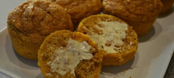 A white platter filled with pumpkin muffins, with one sliced open and spread with butter.