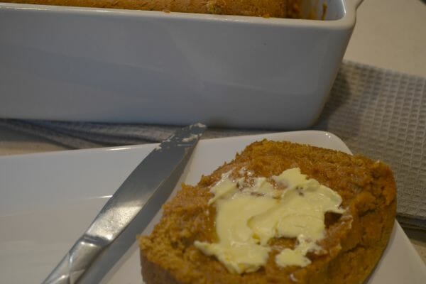 A slice of pumpkin bread on a white plate, spread with butter by a nearby knife.