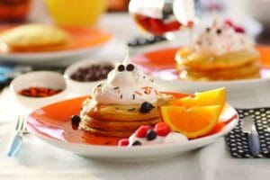 An orange plate with a stack of Pumpkin Pancakes, with whipped cream, berries, sprinkles, and orange slices.