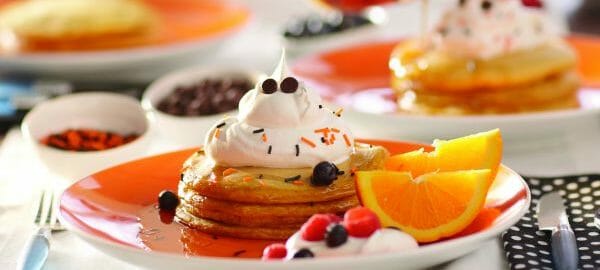 An orange plate with a stack of Pumpkin Pancakes, with whipped cream, berries, sprinkles, and orange slices.