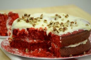 A red and white plate holding a Red Velvet layer cake with Cream Cheese Frosting, topped with chopped nuts.