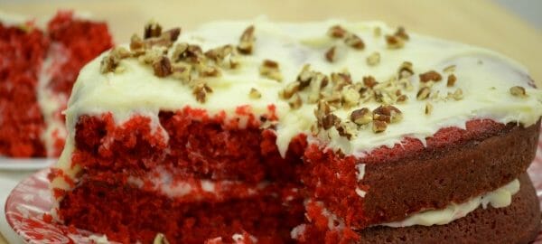 A red and white plate holding a Red Velvet layer cake with Cream Cheese Frosting, topped with chopped nuts.