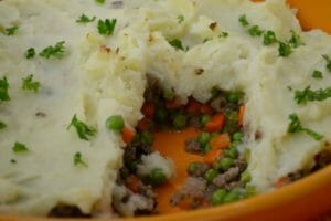 An orange baking dish filled with Shepherd's Pie, with a portion removed to show beef, peas, and carrots.