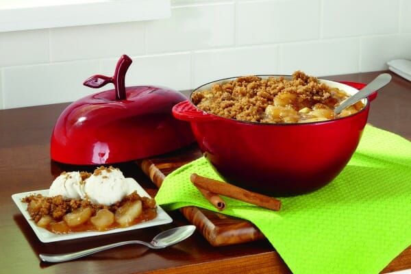 A red apple-shaped covered bowl, filled with Simple Apple Crisp, and a plate with a serving topped with whipped cream.