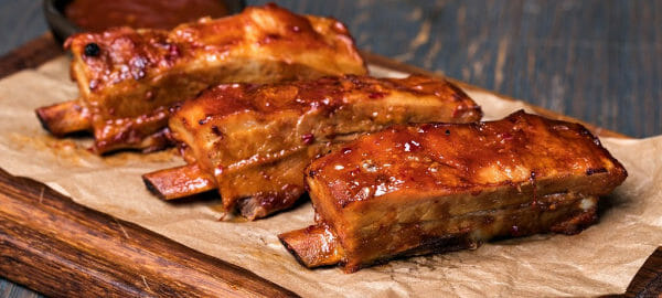 Three Barbeque ribs on a parchment-paper-covered cutting board, with a ramekin of barbeque sauce.