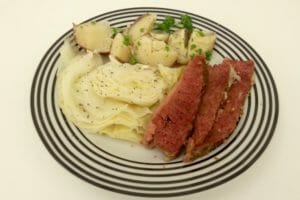 A white plate with black stripes, filled with Slow Cooker Corned Beef, cabbage, and parsley potatoes.
