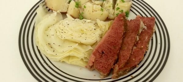 A white plate with black stripes, filled with Slow Cooker Corned Beef, cabbage, and parsley potatoes.