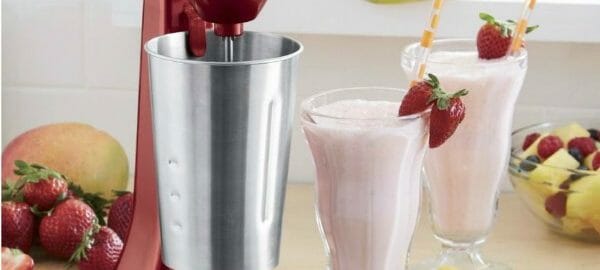 Two strawberry shakes in clear footed glasses, with straws and fresh strawberry garnishes, next to a red blender.
