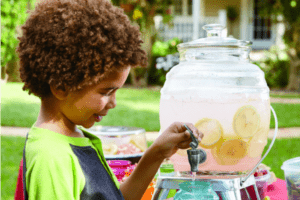 An African-American child outside, pouring a glass of Tart Pink Lemonade from a clear drink dispenser.
