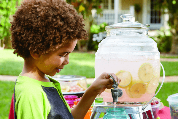 An African-American child outside, pouring a glass of Tart Pink Lemonade from a clear drink dispenser.