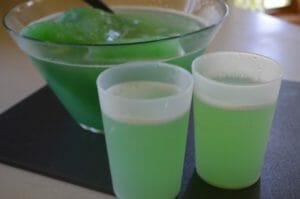 Two frosted glasses filled with green Slushy Punch, next to the filled punch bowl.