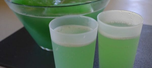 Two frosted glasses filled with green Slushy Punch, next to the filled punch bowl.