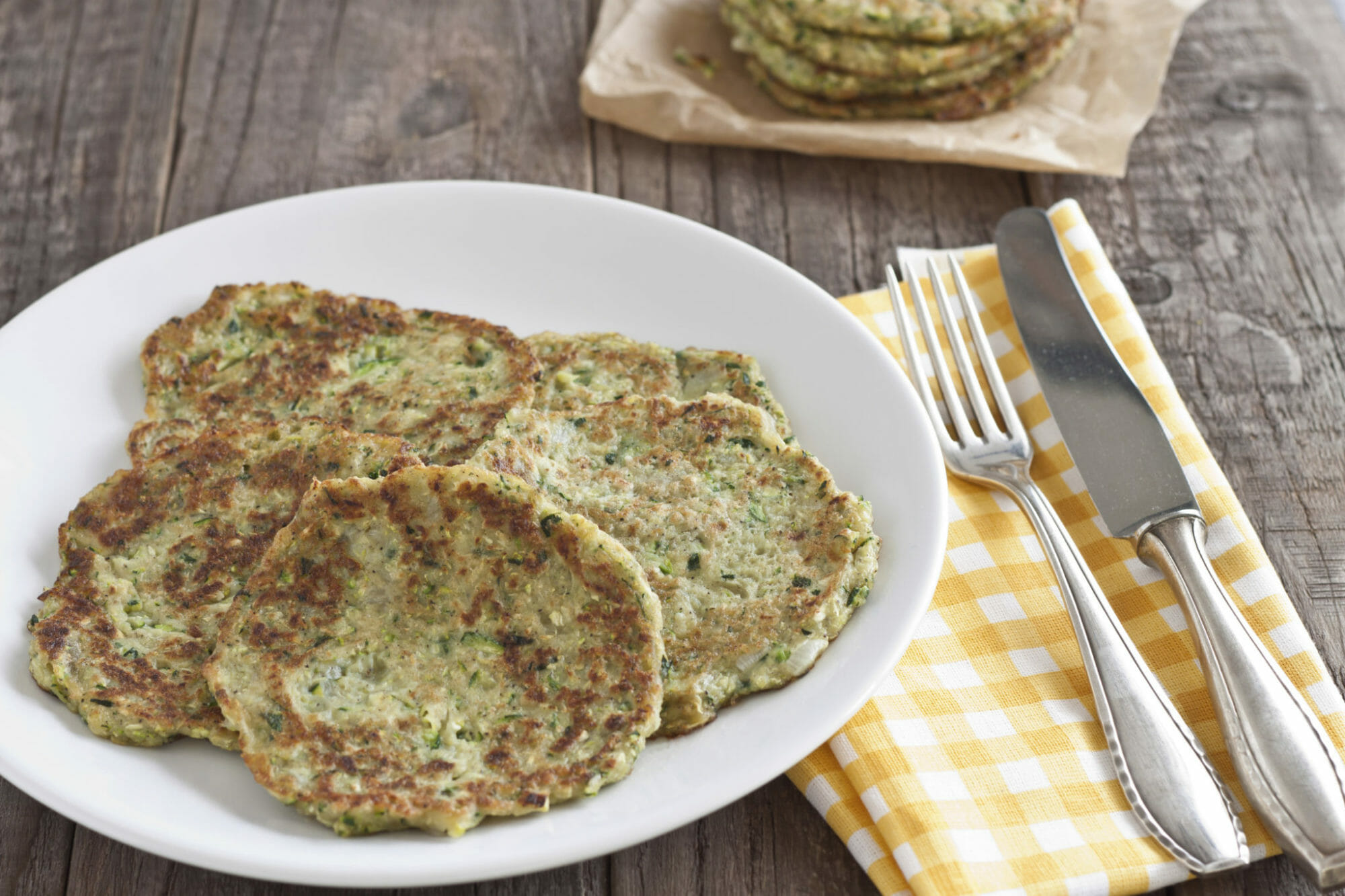 A white plate filled with zucchini patties, next to a fork and knife on a yellow check napkin.