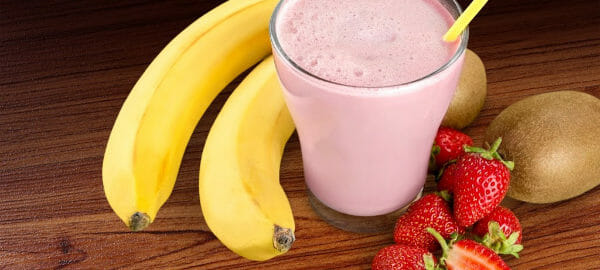 A clear glass filled with a pink Fruit Smoothie and a yellow straw, next to bananas, strawberries, and kiwi.