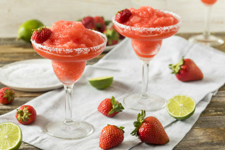 A rustic table with two chilled strawberry margaritas rimmed with coarse sugar and fresh strawberry garnishes.