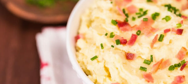 Close-up of a white bowl filled with Bacon Horseradish Mashed Potatoes, topped with chives and bacon pieces.