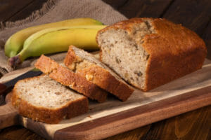 A loaf and three cut slices of Banana Bread on a wooden cutting board, next to two bananas.