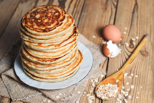 A tall stack of Brown Sugar Oatmeal Pancakes on a white plate, next to a wooden spoon and dried oatmeal flakes.