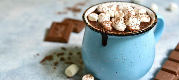 A blue mug filled with Hot Chocolate, topped with cinnamon and mini marshmallows, with a chocolate bar nearby.