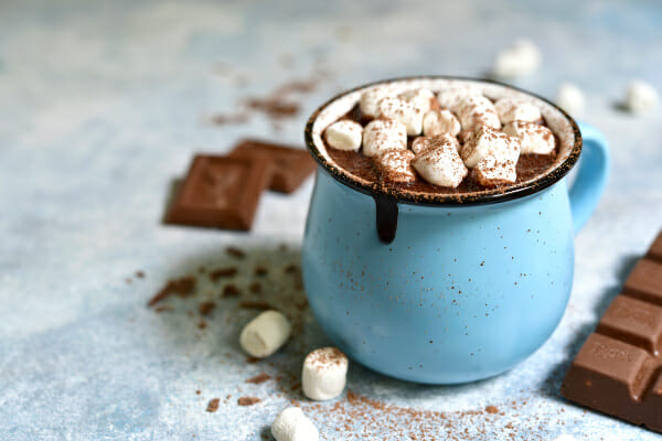 A blue mug filled with Hot Chocolate, topped with cinnamon and mini marshmallows, with a chocolate bar nearby.