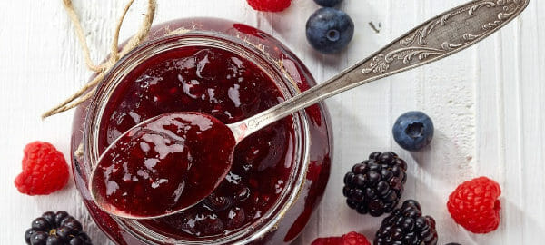 A jelly jar and a spoon filled with jam, surrounded with blueberries, raspberries, and blackberries.