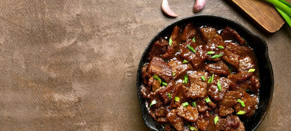 A black cast iron skillet filled with glazed Mongolian Beef, with green onions on a cutting board nearby.