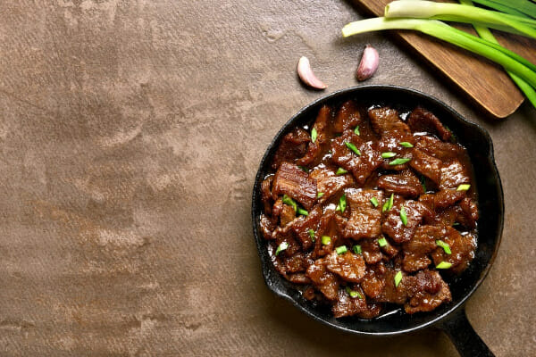 A black cast iron skillet filled with glazed Mongolian Beef, with green onions on a cutting board nearby.