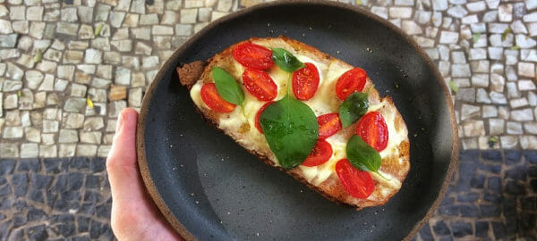 A hand holding a black spattered plate with Caprese Toast topped with cherry tomatoes and basil.