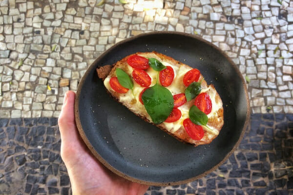 A hand holding a black spattered plate with Caprese Toast topped with cherry tomatoes and basil.