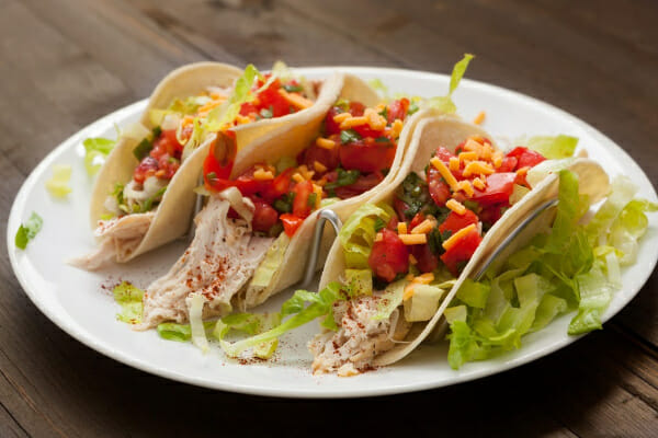 A white plate filled with chicken tacos topped with tomato, lettuce and shredded cheese.