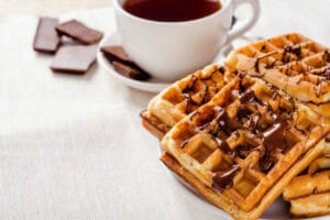 A white plate stacked with waffles topped with drizzles of chocolate, next to a white cup of hot chocolate.