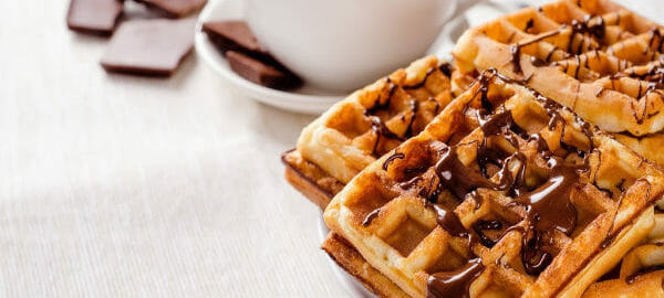 A white plate stacked with waffles topped with drizzles of chocolate, next to a white cup of hot chocolate.