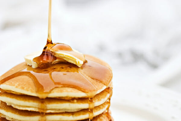 Maple syrup being poured on a stack of fluffy pancakes topped with butter pats.