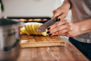 A woman's hand cutting potato slices with a sharp knife for fried potatoes.