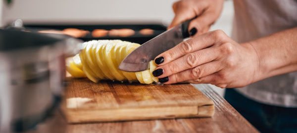A woman's hand cutting potato slices with a sharp knife for fried potatoes.
