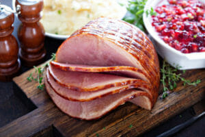 A ham with four slices on a wooden cutting board next to a white bowl filled with cranberry sauce.