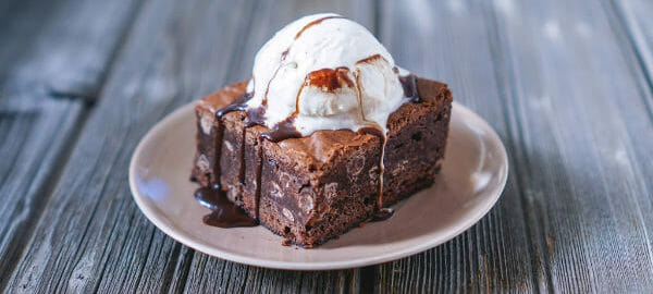 A white dessert plate with a slice of Hot Fudge Cake, topped with chocolate drizzles and vanilla ice cream.