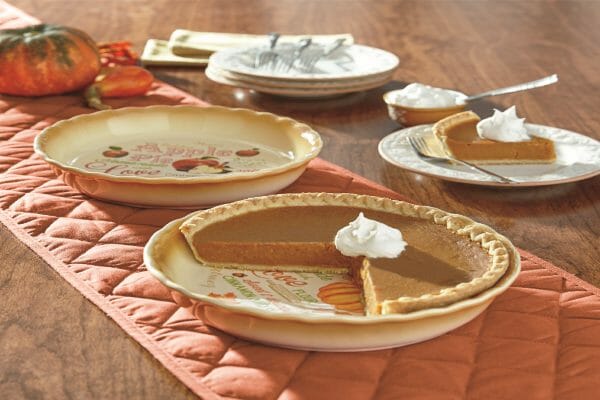 An orange table runner with two pie plates, one filled with pumpkin pie, and a slice topped with whipped cream.