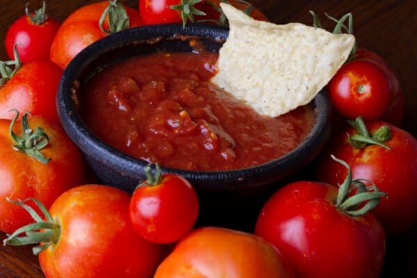 A black bowl filled with restaurant-style salsa and a tortilla chip, surrounded by red tomatoes.