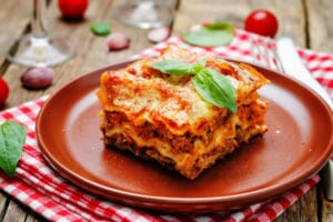 A rust-colored plate with a serving of slow cooker Lasagna topped with a sprig of basil, on a rustic picnic table.