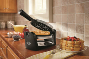 A black Ginny's brand waffle iron on a kitchen counter, with a stack of waffles on a plate, topped with fresh fruit.