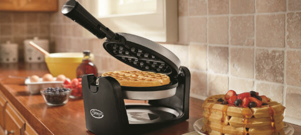 A black Ginny's brand waffle iron on a kitchen counter, with a stack of waffles on a plate, topped with fresh fruit.