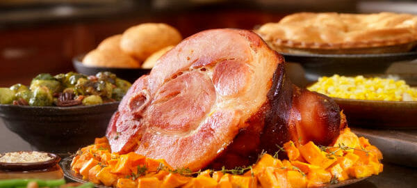 A Holiday table filled with a ham on a bed of squash, with bowls of vegetable dishes, rolls, and a pie.