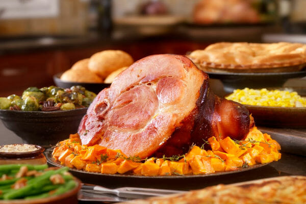 A Holiday table filled with a ham on a bed of squash, with bowls of vegetable dishes, rolls, and a pie.